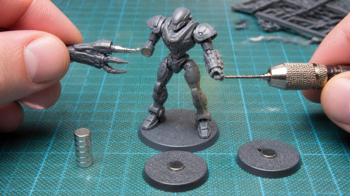Hands drilling a robot miniature with a pin vice while holding a magnetised weapon arm, with disc magnets and magnetised bases on a cutting mat