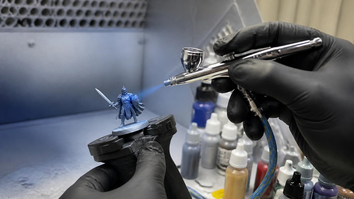 Gloved hands airbrushing blue paint onto a knight miniature on a painting handle inside a spray booth