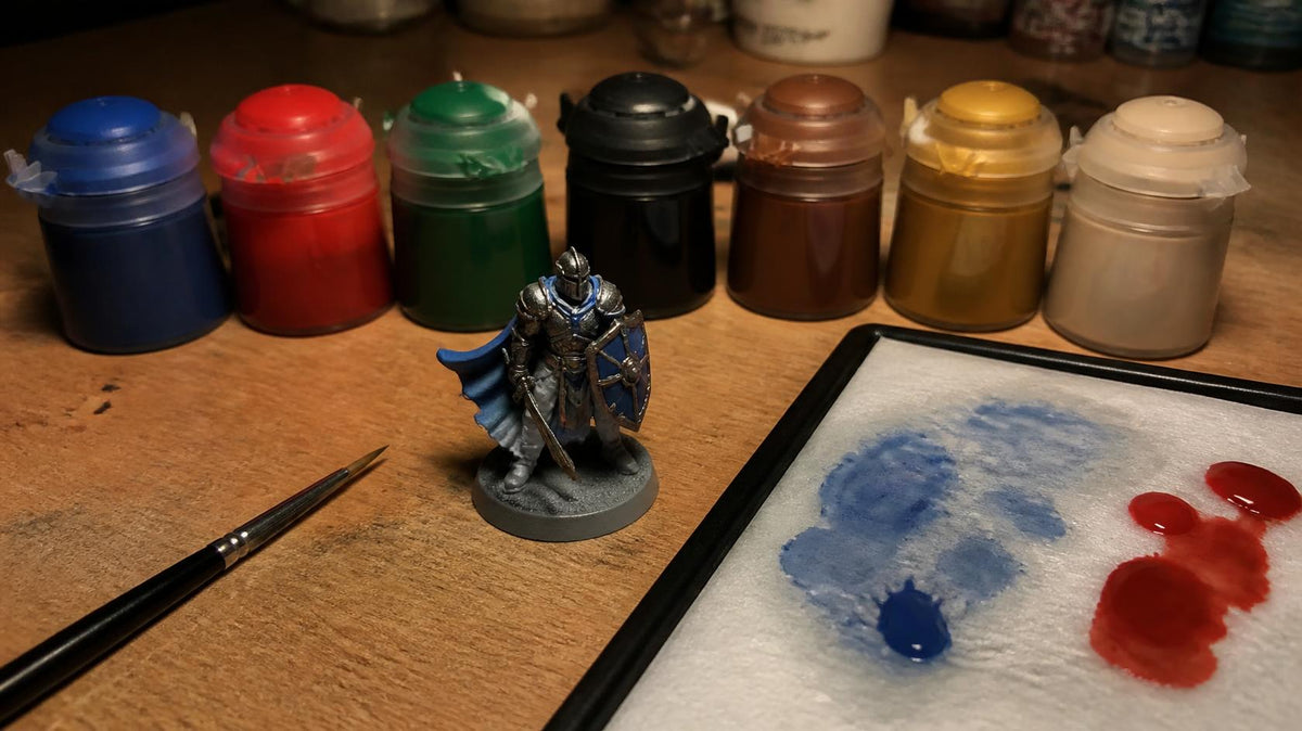 Row of hobby paint pots behind a part-painted knight miniature, a detail brush and a wet palette with blue and red paint