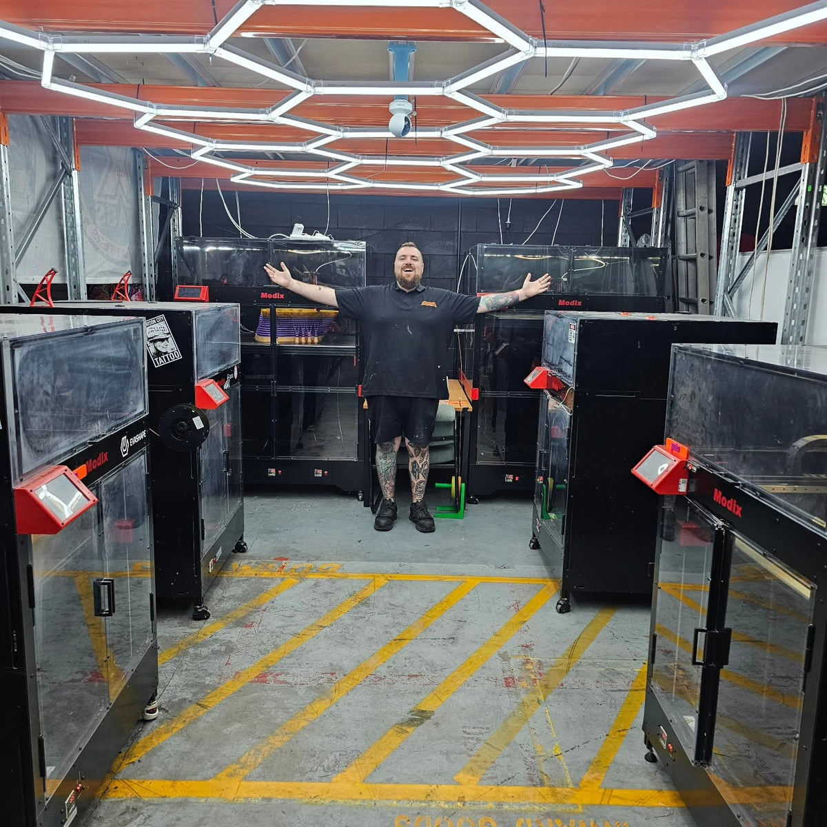 Zak standing with arms outstretched among the large-format Modix 3D printers in the Rising Sun FPV workshop in Kirwan